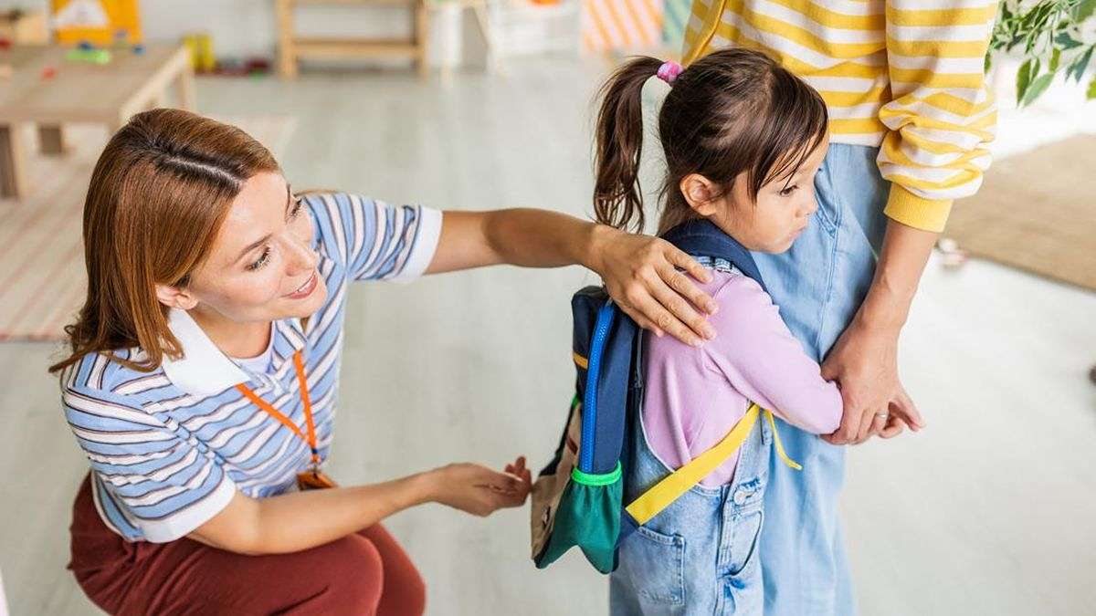 Latih 5 Kalimat Agar Anak Berani Bicara demi Masa Depan Si Kecil 53 Guru sekolah menenangkan seorang anak perempuan yang memegang tas punggung dan berlindung di belakang orang tuanya