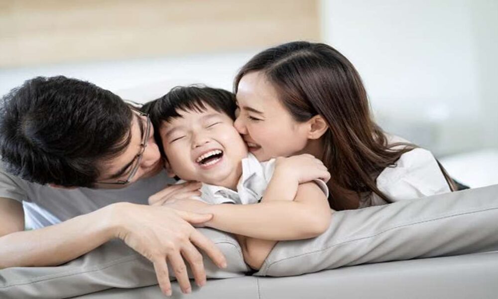 Seorang anak tertawa ceria saat dipeluk dan dicium oleh ayah dan ibunya di sofa