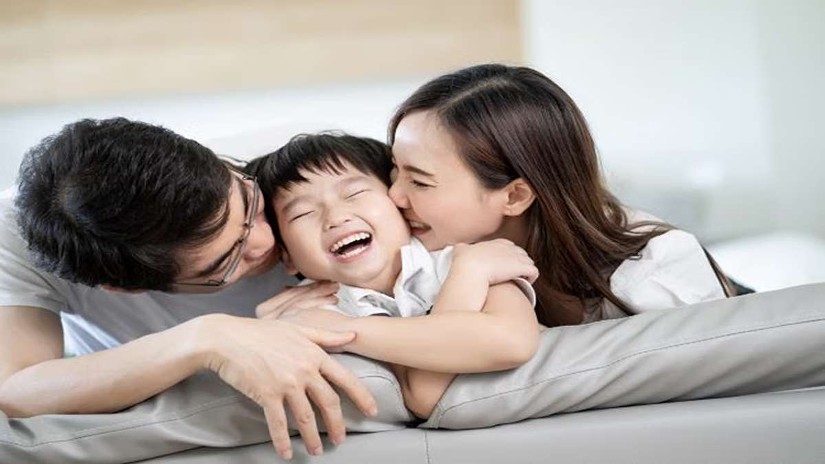Seorang anak tertawa ceria saat dipeluk dan dicium oleh ayah dan ibunya di sofa